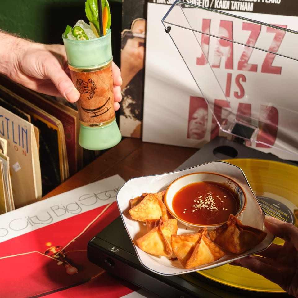 Hand holds a green cocktail garnished with jalapeño and citrus beside fried dumplings with sesame dipping sauce in a colorful restaurant setting.