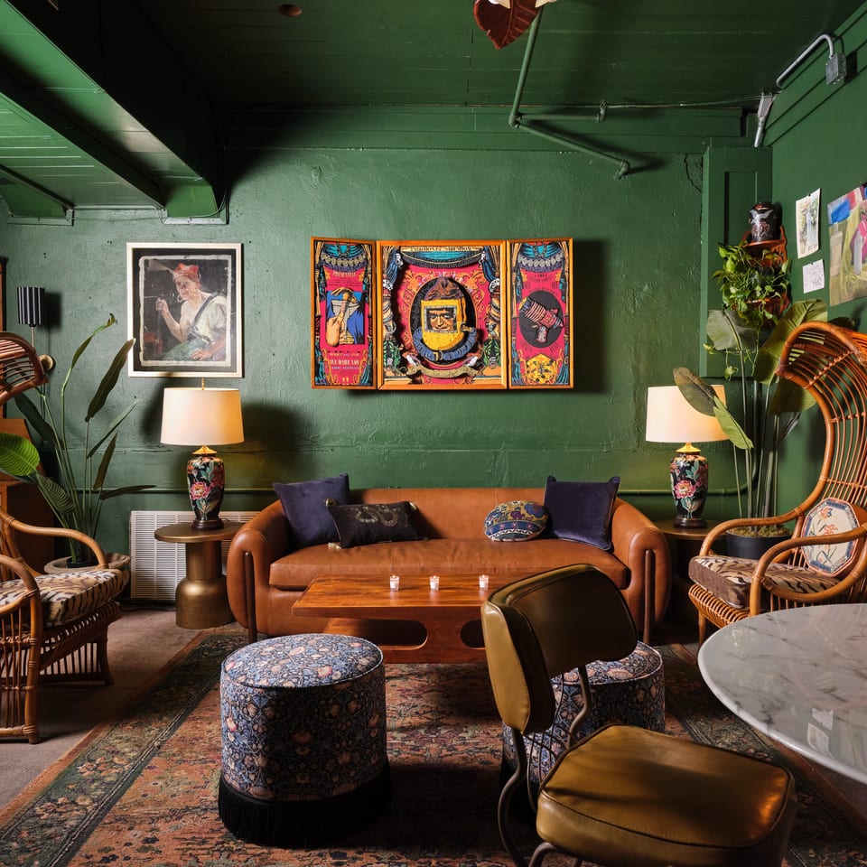 Eclectic restaurant lounge with green walls, rattan chairs, leather sofa, patterned rugs, plants, lamps, and colorful artwork.