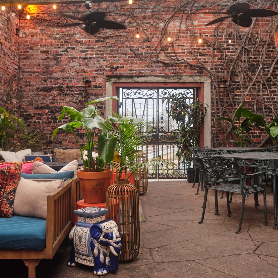 Cozy restaurant courtyard with exposed brick walls, string lights, ceiling fans, tropical plants, colorful cushions, and ornate wrought-iron tables.