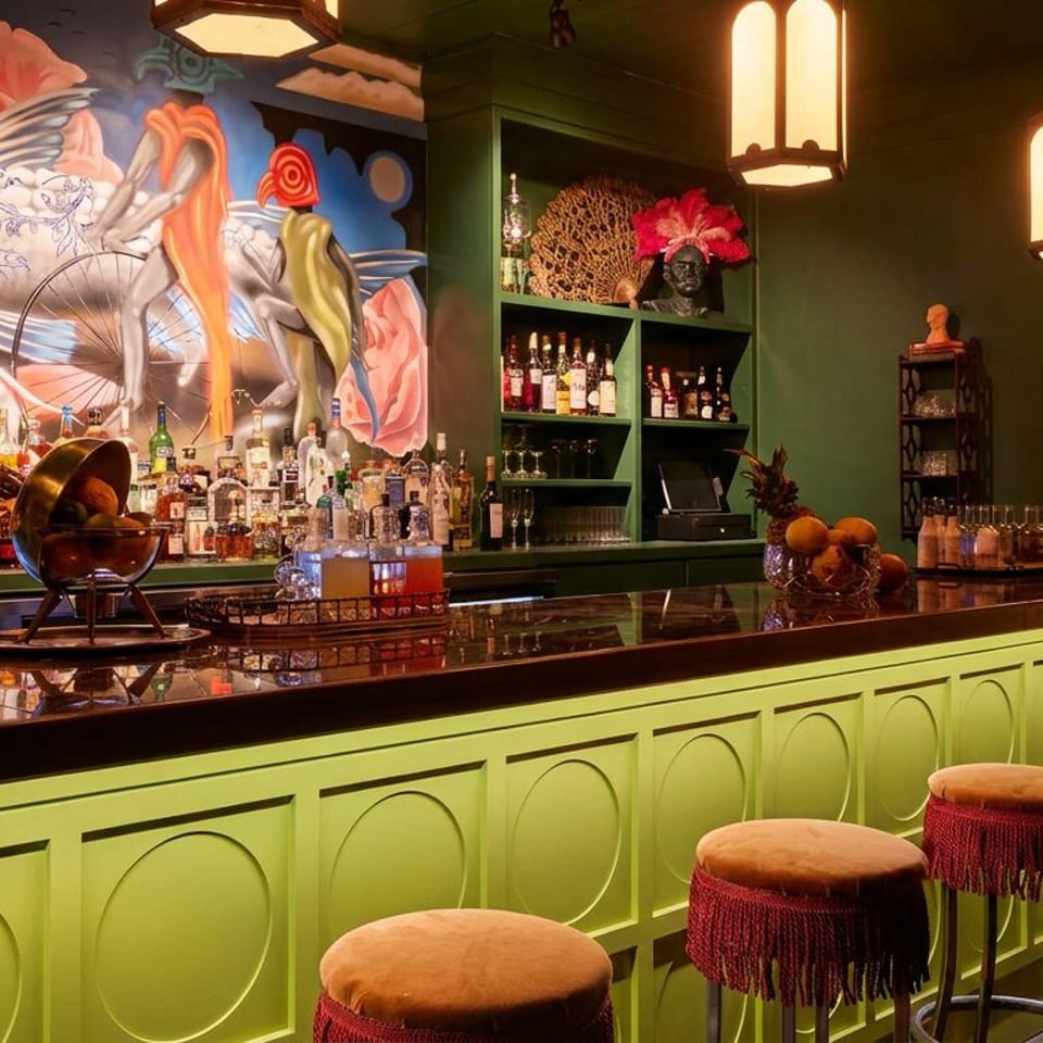 A vibrant bar with a green counter, red-fringed stools, and a colorful mural behind liquor bottles.