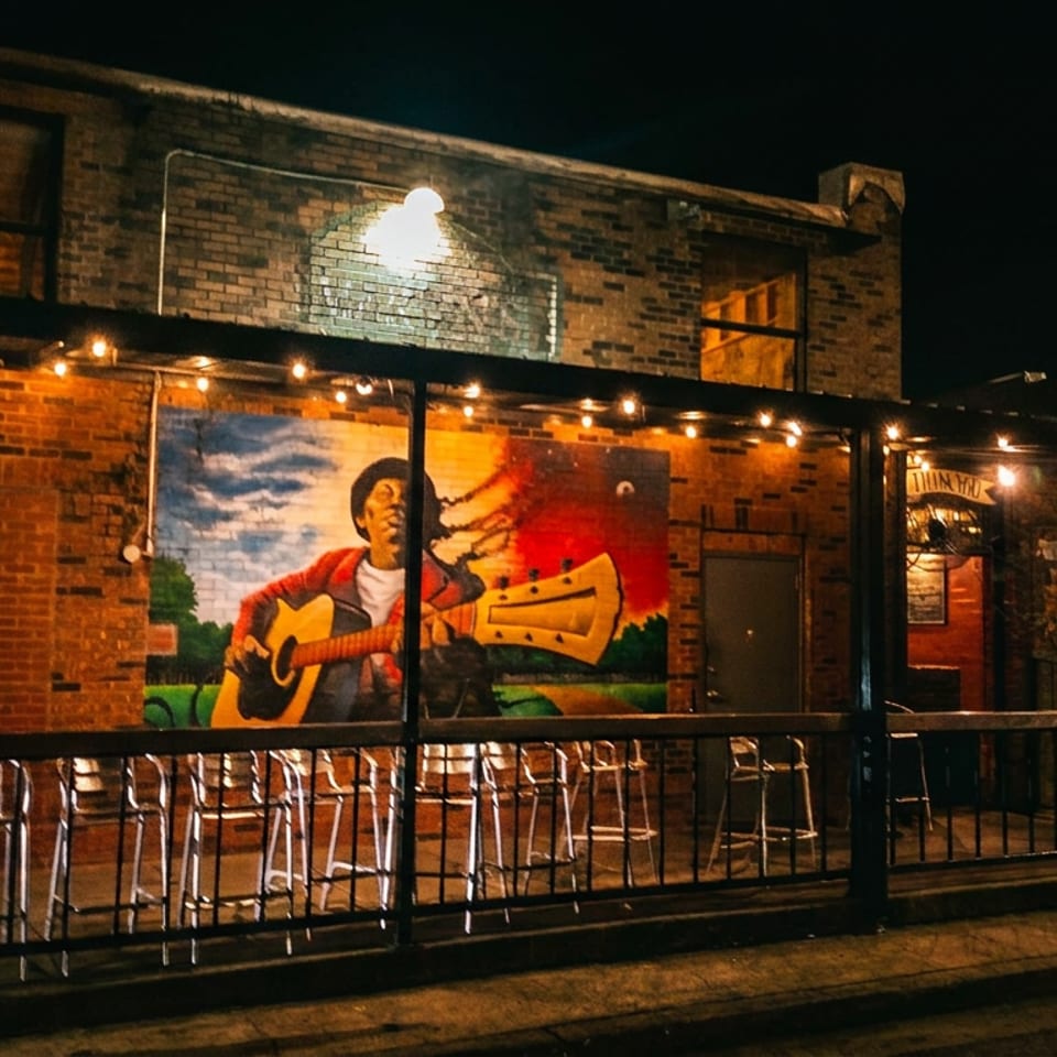 Night view of a brick building with string lights, a mural of a man playing guitar, and an outdoor patio.