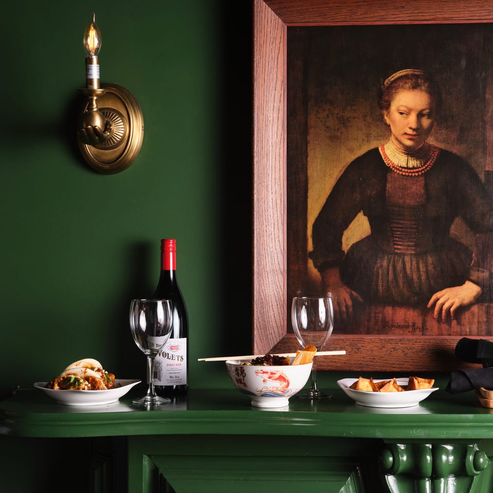 Restaurant table displays Asian dishes, wine, chopsticks, and napkins beneath a wall sconce and framed portrait.