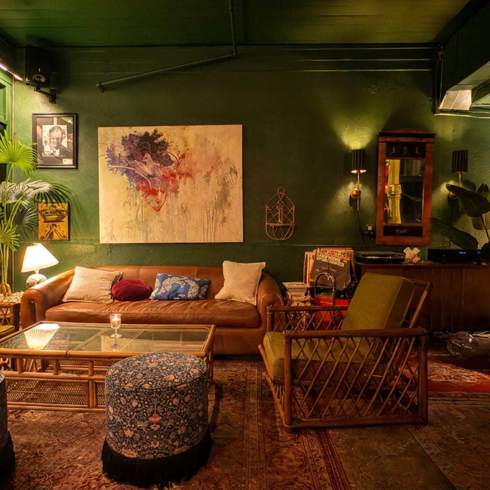 A cozy, dimly lit lounge with green walls, leather sofa, rattan chairs, and eclectic decor.