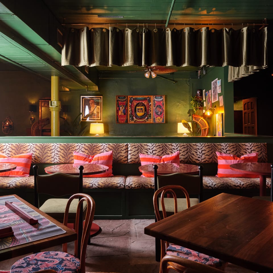 Warmly lit restaurant dining room with patterned banquette seating, colorful cushions, wooden tables, artwork, and a vintage patterned rug.