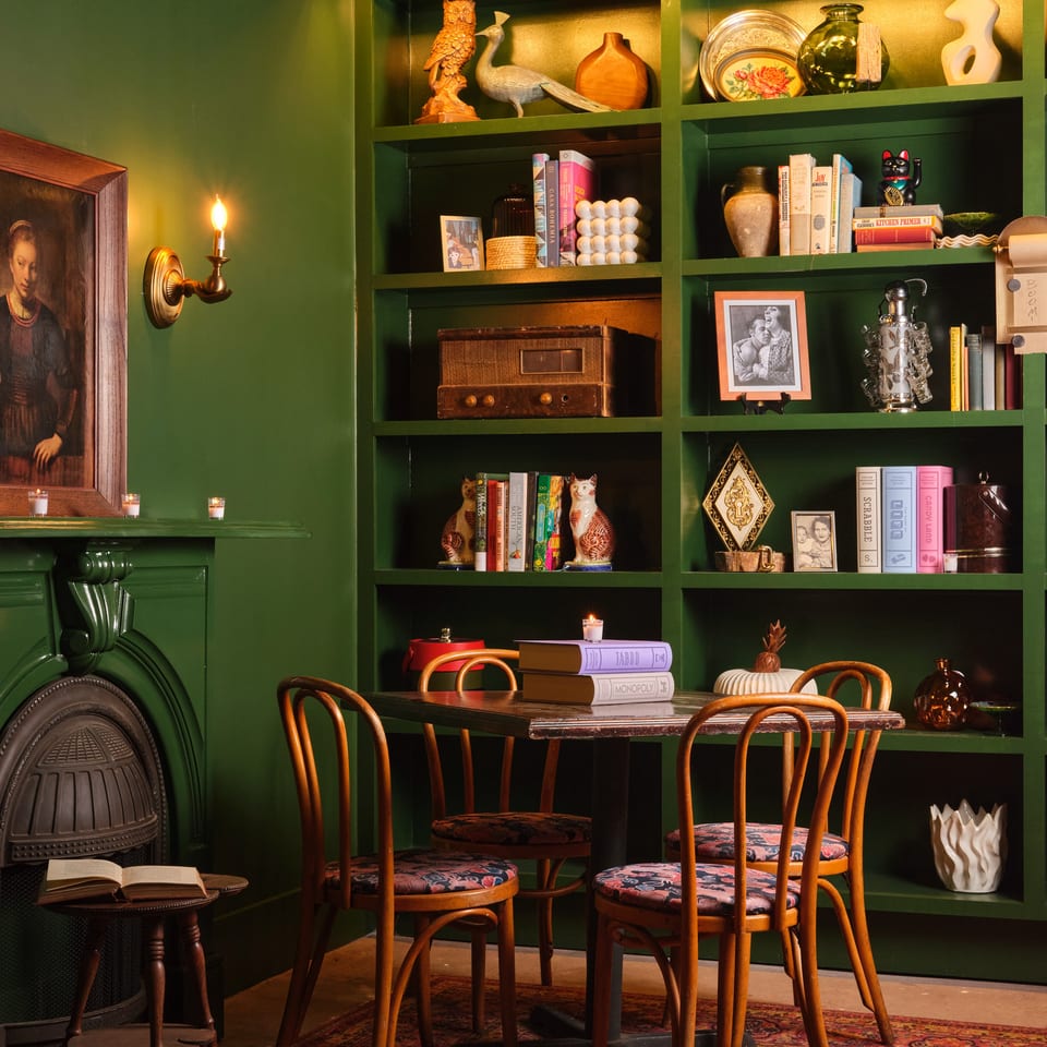 Moody restaurant interior with dark green shelves displaying books, vintage objects, candles, and a dining table with patterned chairs.