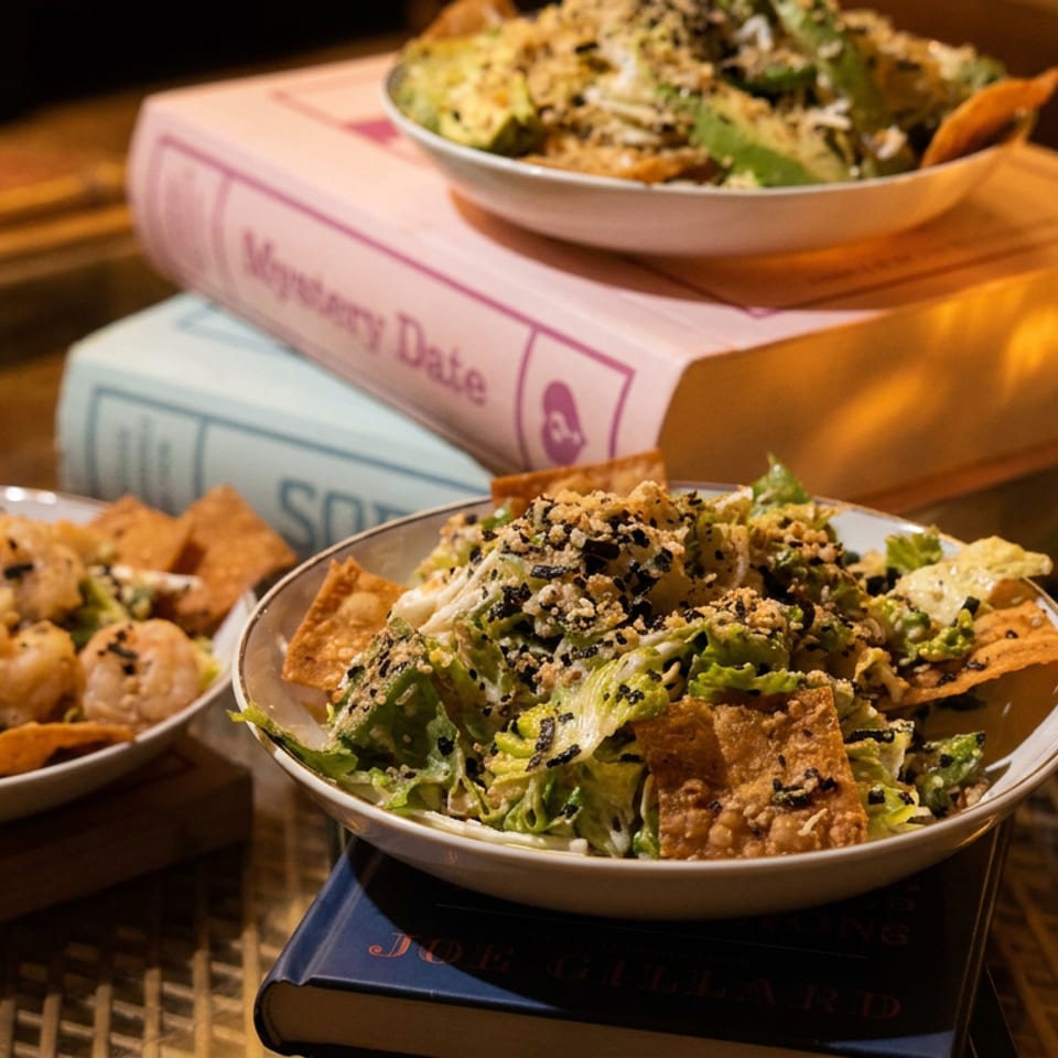 Three bowls of fresh salad with crispy toppings and shrimp, alongside books and a candlelit glass.