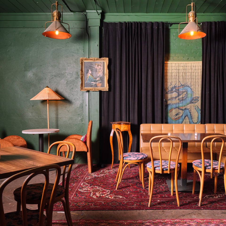 Eclectic restaurant dining room with green walls, vintage lamps, dark curtains, patterned rugs, wooden tables, and tan banquette seating.